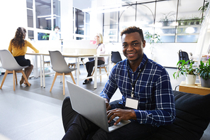 Cheerful handsome young black manager wearing conference badge on neck sitting in bean bag and typing on laptop while working in comfortable office