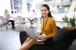 Positive beautiful young woman with badge sitting in comfortable bean bag and smiling at camera, while typing on laptop, she working in cozy office