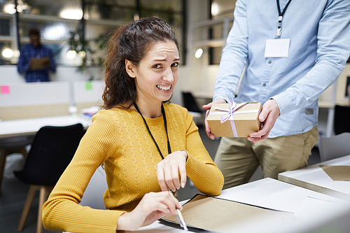 Displeased frowning attractive young lady in sweater disgusted by colleagues gift sitting at table and  while working on project design