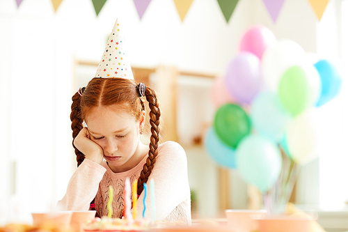 Portrait of sad red haired girl sitting alone at party table in decorated room, copy space