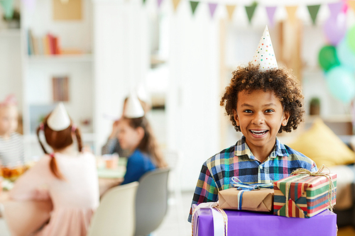 Portrait of happy African-American boy holding gift boxes posing during birthday party with friends, copy space