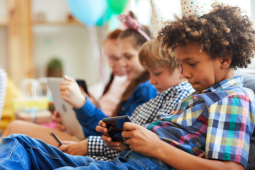 Side view at group of children using smartphones sitting in row at birthday party, copy space