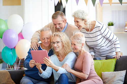 Smiling excited curly-haired lady in glasses holding tablet and taking congrats from friends online using telecommunication app, her friends supporting her and being part of internet conversation