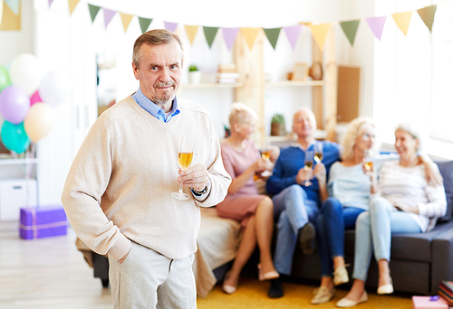 Portrait of content handsome elderly man with beard wearing casual sweater drinking champagne from flute at birthday party while his friends chatting in background
