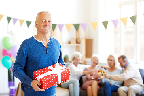 Portrait of content handsome birthday man in blue sweater standing in living room decorated for birthday party and holding gift box while his friends clinking flutes in background.