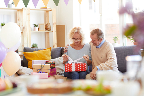 Content attractive curly-haired lady in glasses embraced by husband sitting in sofa in living room and opening birthday gifts