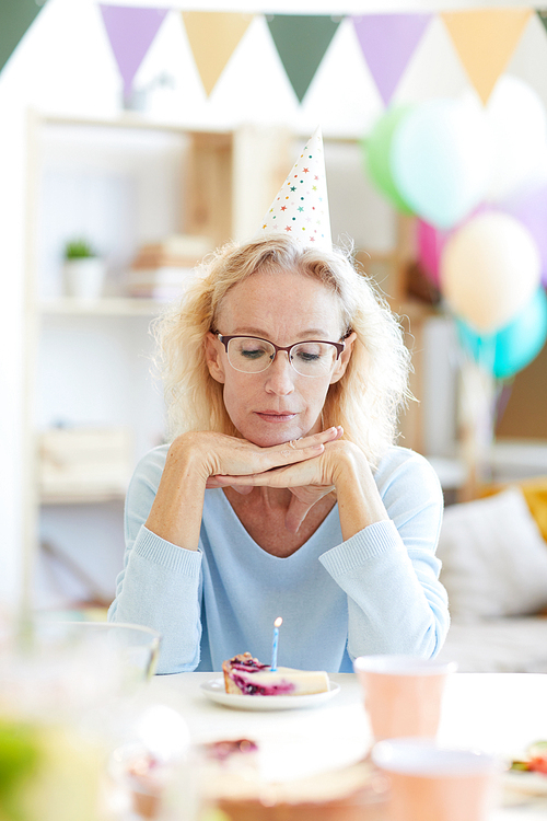 Depressed beautiful mature lady in party hat sitting at table and leaning on hands while thinking about birthday wish
