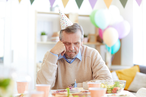 Lonely bearded mature man in party hat sitting at table in decorated living room and looking at birthday cake with candle