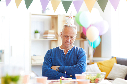 Serious unhappy mature man in party hat sitting at table and keeping arms crossed while making wish at own birthday