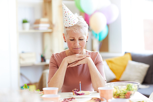 Serious pensive elderly woman in party hat sitting at table and looking at birthday cake while celebrating birthday alone