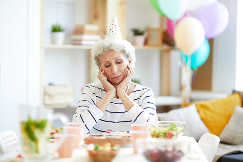 Serious unhappy retired gray-haired lady in party hat sitting at dining table full of dishes and looking at birthday cake with candle while having birthday party alone.