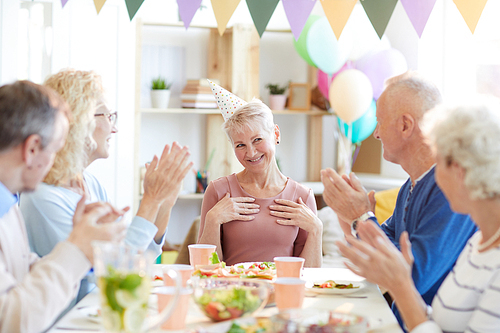 Group of positive mature friends sitting at dining table in living room and clapping hands for birthday woman while congratulating her together