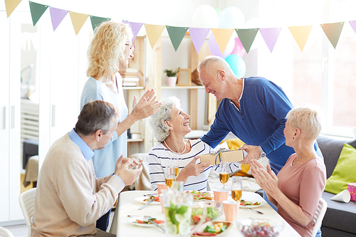 Group of positive excited people celebrating birthday of retired pretty woman at home, handsome man giving gift to birthday woman