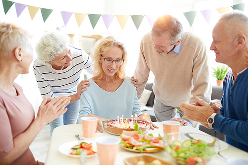 Smiling excited mature birthday woman with curly hair sitting at table and thinking about wish while preparing to blow candles on sweet cake