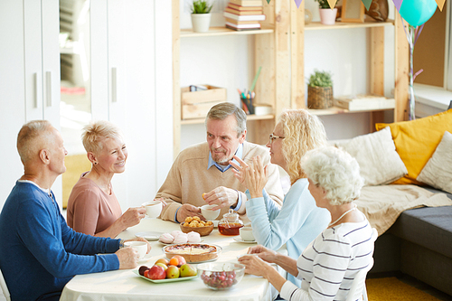 Group of positive mature friends in casual clothing sitting at table and drinking tea with sweets while chatting during party