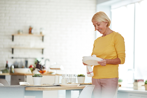 Positive attractive mature woman with short blond hair standing at dining table and wiping plate with dish towel while preparing for dinner party at home