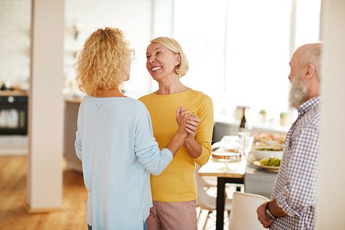 Cheerful attractive mature lady friends in casual outfits excited about gathering holding hands and dancing together in flat studio while man looking at them