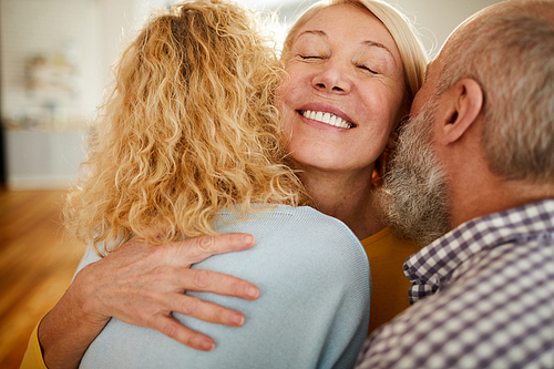 Happy excited mature woman with closed eyes embracing friends while welcoming them to home