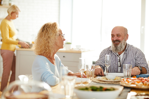 Jolly excited mature friends in casual clothing sitting at table and talking to each other while waiting for dinner, woman cooking in background