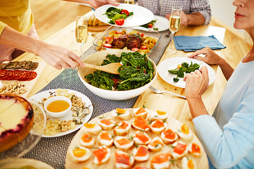 Close-up of unrecognizable woman standing at dining table full of appetizers and dishes and putting vegetable salad on plate at dinner