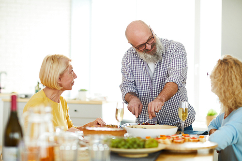 Content handsome mature bearded man in glasses standing at table and cutting meat for guests while talking to charming ladies at home