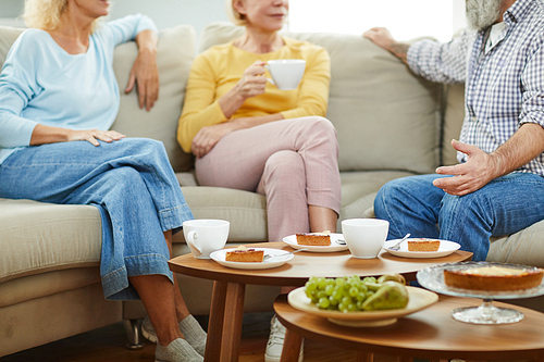 Close-up of mature friends in casual outfits sitting on sofa and drinking tea while chatting during tea party