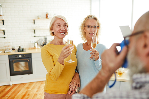 Cheerful attractive mature ladies in casual clothing standing in kitchen room and holding champagne flutes while posing for camera