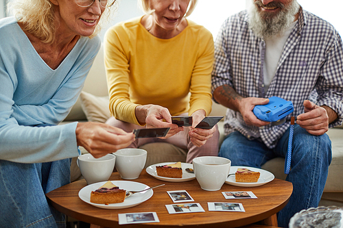 Close-up of mature friends sitting at coffee table in shape of leaf and viewing photos made on instant camera while drinking tea and eating cake.