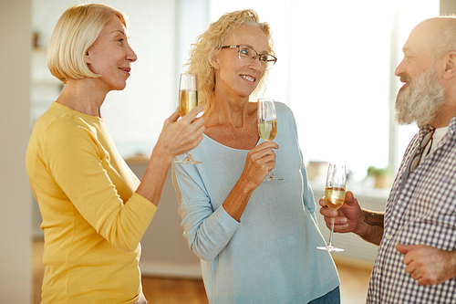 Group of cheerful mature friends in casual clothing drinking champagne and chatting together while having house party