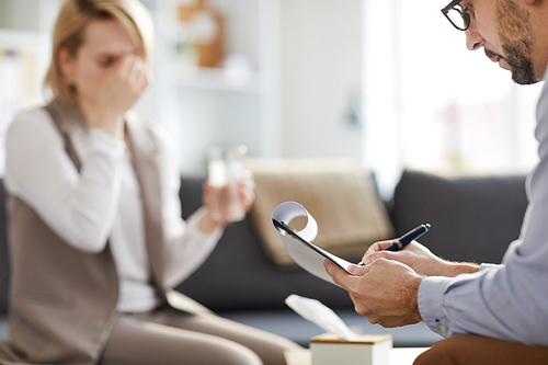 Young confident counselor holding document and making notes while communicating woth one of troubled patients