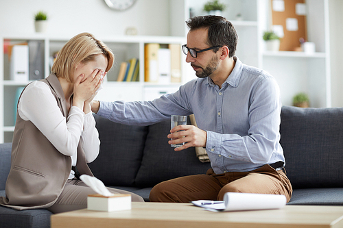 Professional counselor in eyeglasses offering glass of water to his crying patient while both sitting on couch