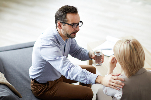 Contemporary professional psychologist with glasss of water comforting his patient during session