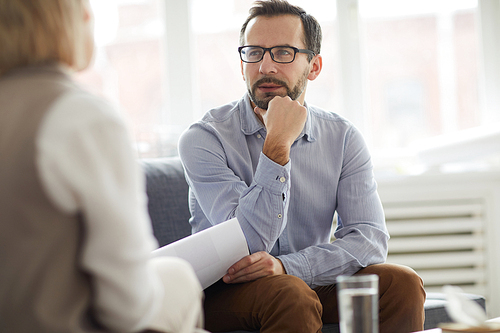 Pensive counselor with paper sitting on couch in front of patient and listening to her explanation of problem