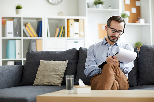 Young professional counselor making notes in document while sitting on couch in his office after talk with client