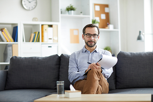 Young successful pychologist looking at you while sitting on couch in front of camera in his office