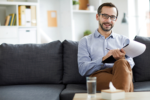 Successful cheerful psychologist with document making notes while sitting on couch in front of camera