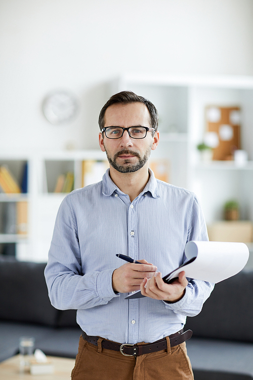 Young serious man in shirt, pants and eyeglasses standing in front of camera while making notes