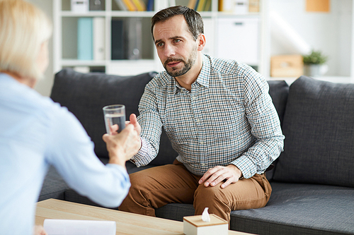 Young troubled man taking glass of water from his counselor while having discussion of his problems