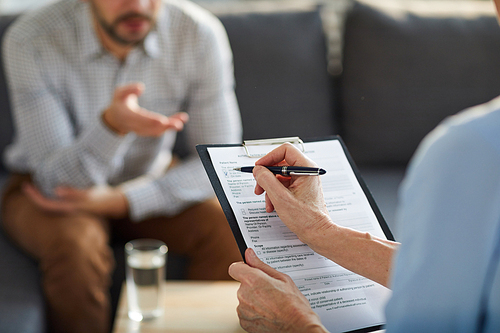 Hand of mature professional counselor with pen over medical document putting tick during conversation with patient