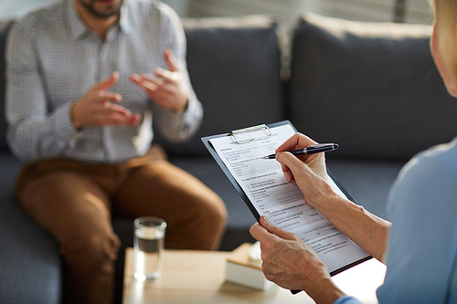 Mature professional counselor filling in document while having discussion with patient in trouble