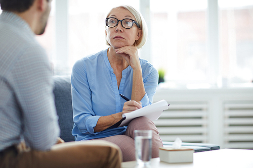 Serious blonde mature counselor in eyeglasses listening to patient telling his story and explaining the problem