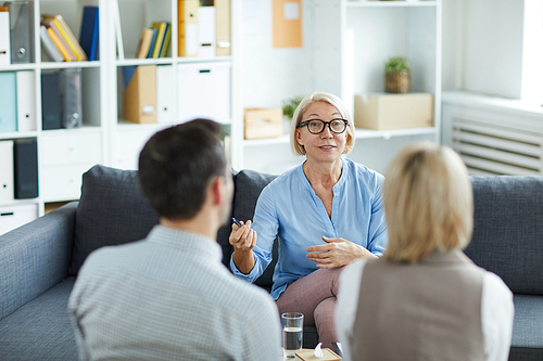 Mature blonde female psychologist sitting in front of young troubled couple and consulting them in office
