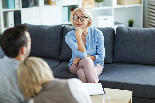 Confident mature psychologist in smart casual sitting on couch in front of young troubled couple during session