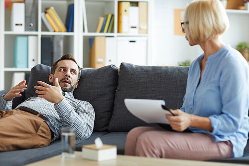 Young patient of counselor lying on couch, looking at mature professional and explaining his problem