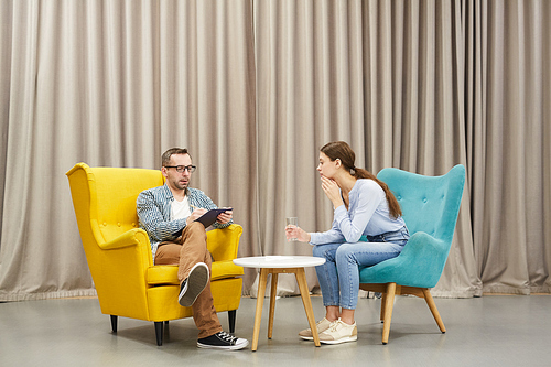 Full length portrait of young woman talking to mature psychologist in therapy session sitting on design chairs against drapery, copy space