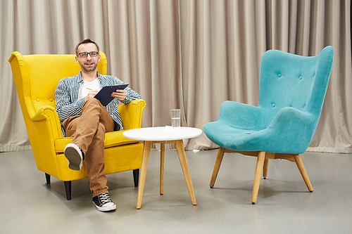 Full length portrait of mature psychologist holding clipboard  posing  while  sitting on design chair against drapery, copy space