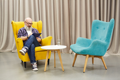 Full length portrait of senior psychologist  holding clipboard sitting in design chair against drapery, copy space