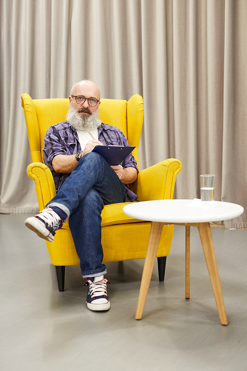 Full length portrait of senior psychologist holding clipboard  posing  while  sitting on design chair against drapery