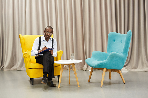 Full length portrait of African-American man holding clipboard  posing  while  sitting on  design chair against drapery