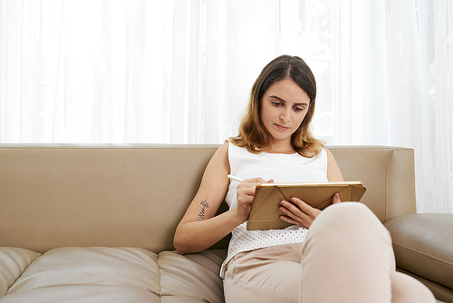 Pretty young woman sitting on sofa and home and drawing on tablet computer